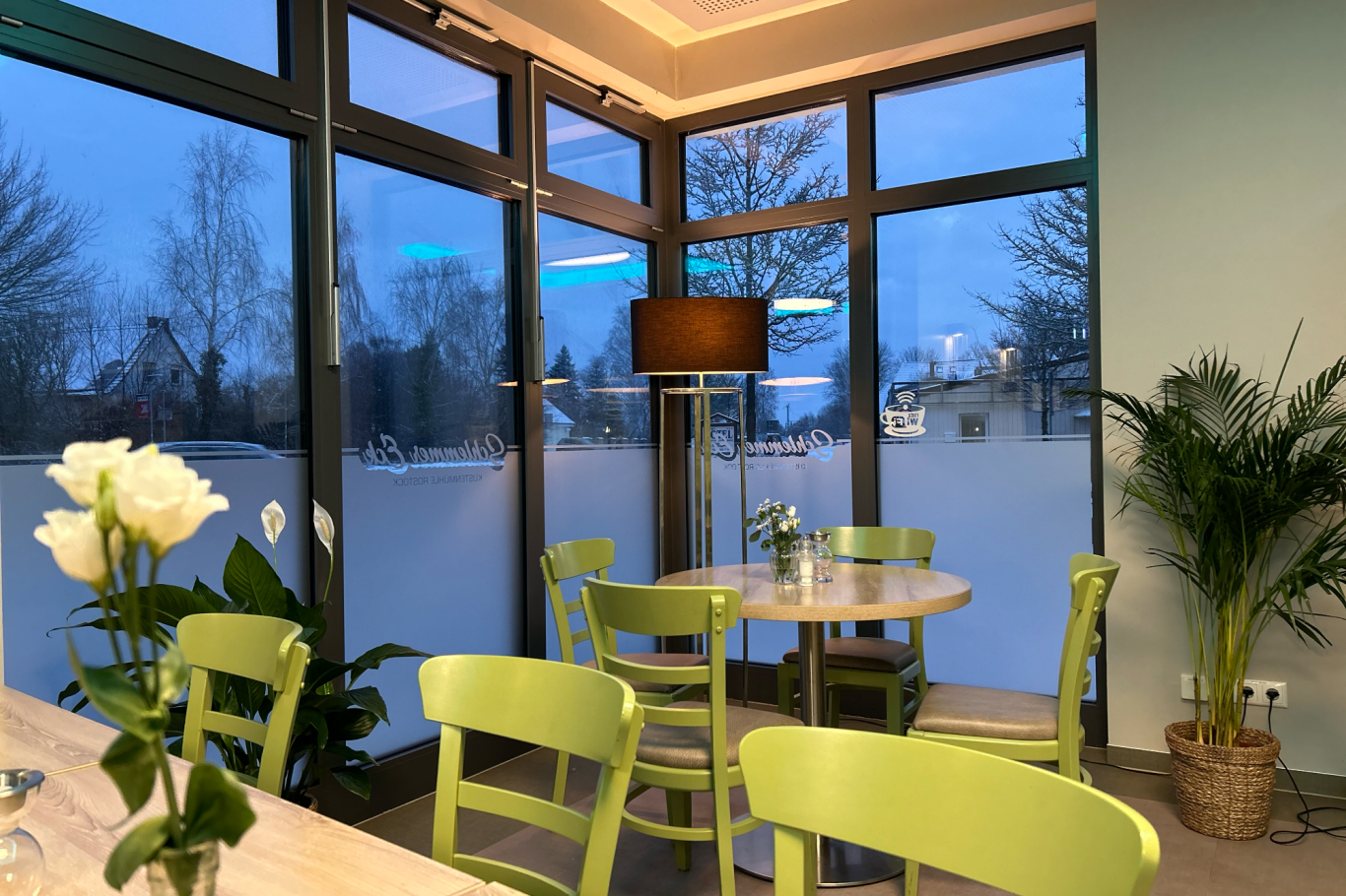 Auf dem Bild ist die Sitzecke in der Kantine zu sehen, in welche angenehm grüne Stühle, ein Tisch und diverse Grünplatzen stehen.